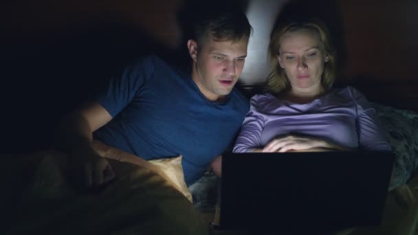 Par, man och kvinna, titta på en film på en bärbar dator på en säng i sovrummet innan sängen. Titta på en spännande film, ser publiken med intresse, upplever olika känslor. — Stockvideo