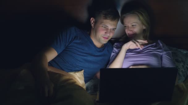 Paar, man en vrouw, lettend op een film op een laptop op een bed in de slaapkamer voor het slapen. kijken naar een komedie-film, lachen het publiek. — Stockvideo