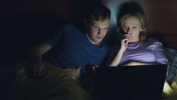 Par, man och kvinna, titta på en film på en bärbar dator på en säng i sovrummet innan sängen. Titta på en spännande film, ser publiken med intresse, upplever olika känslor. — Stockvideo