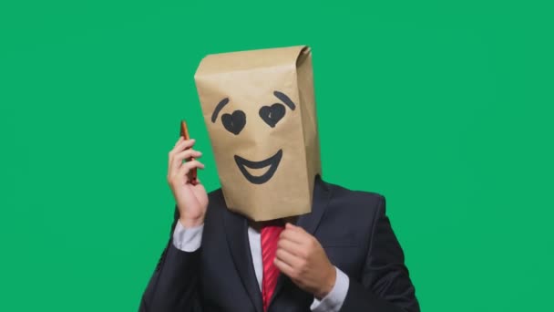 Concepto de emociones, gestos. un hombre con bolsas de papel en la cabeza, con un emoticono pintado, sonrisa, alegría, amor. hablando por un teléfono celular — Vídeos de Stock