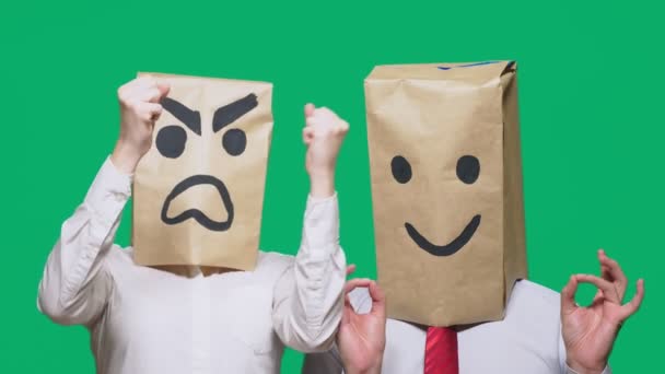 The concept of emotions and gestures. Two people in paper bags on the head with painted smileys. Aggressive smiley swears. The second smiles at him. — Stock Video