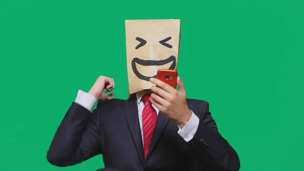 Αντίληψη των συναισθημάτων, χειρονομίες. ένας άνθρωπος με χάρτινες σακούλες στο κεφάλι του, με ζωγραφισμένα φατσούλα, χαμόγελο, χαρά. μιλάμε για ένα κινητό τηλέφωνο — Φωτογραφία Αρχείου