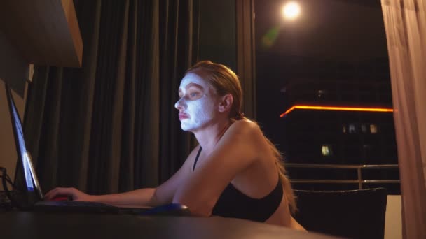 Ung kvinna med kosmetiska mask på ansikte arbeta med laptop hemma på kvällen. naturlighet — Stockvideo