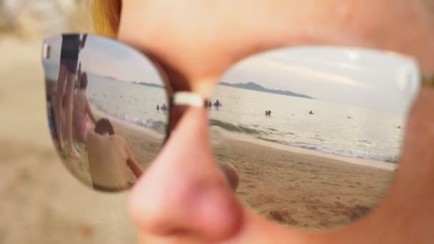 Gros plan d'une femme heureuse portant des lunettes de soleil. La plage avec les personnes au repos se reflète dans les lunettes . — Video
