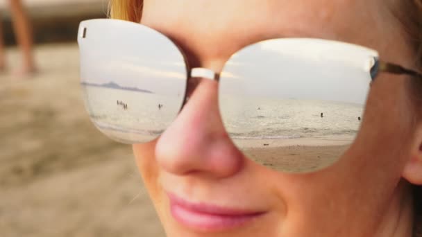 Γκρο πλαν το πρόσωπό του μια ευτυχισμένη γυναίκα σε γυαλιά ηλίου. Η παραλία με τα στηργμένος άτομα αντανακλάται στα γυαλιά. — Αρχείο Βίντεο