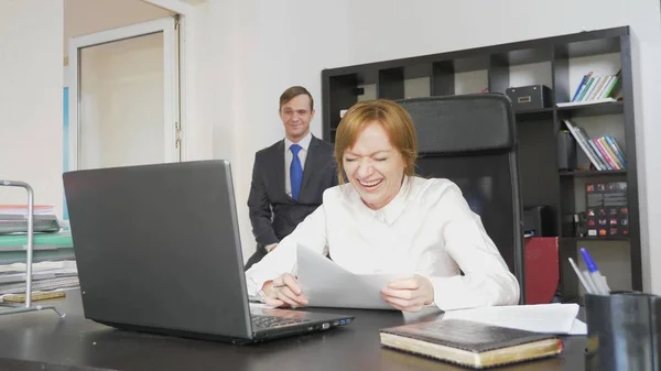 Zwei Büroangestellte sitzen am Schreibtisch, eine Frau arbeitet am Computer, ein Mann ist in der Nähe. sie lachen. — Stockfoto