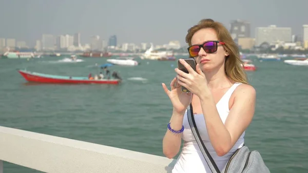 Vrouw in zonnebril, gebruikt haar smartphone terwijl staande op de pier. achtergrond vervagen, — Stockfoto