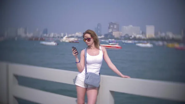 Mujer en gafas de sol, utiliza su teléfono inteligente mientras está de pie en el muelle. desenfoque de fondo , — Foto de Stock