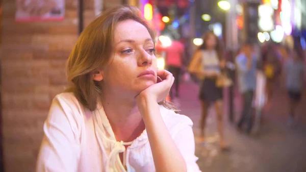 Mujer joven y hermosa sentada en la calle en un café o terraza. Una mujer observa a los transeúntes caminando por una calle peatonal por la noche . — Foto de Stock