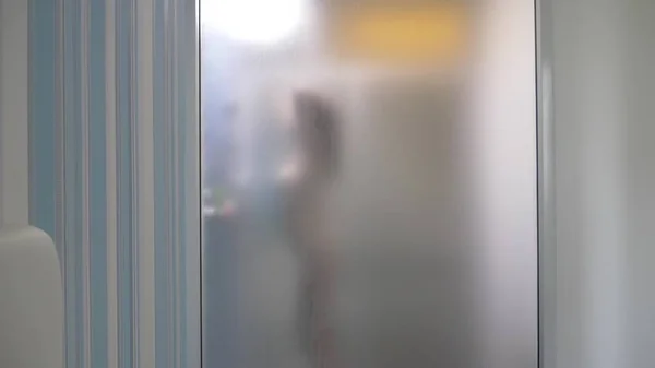 Mujer detrás de vidrio borroso. Chica preparando tomar ducha. Mujer en el baño. un hombre mira como una mujer toma una ducha a través de una pared de vidrio en la ducha . — Foto de Stock