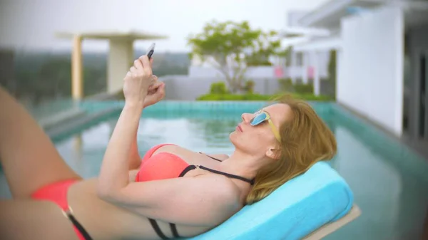 Una mujer en bikini rosa junto a la piscina en el techo, una chica tomando el sol en una tumbona y usando el teléfono — Foto de Stock