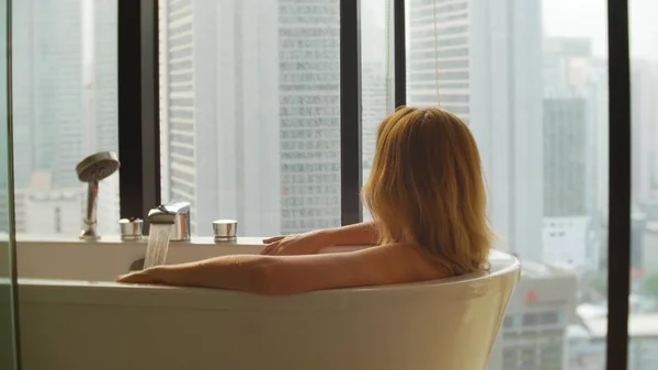 Женщина наслаждается релаксационной ванной в роскошной ванной с окном. Концепция ухода за образом жизни и красотой. вид из окна на небоскребы . — стоковое фото