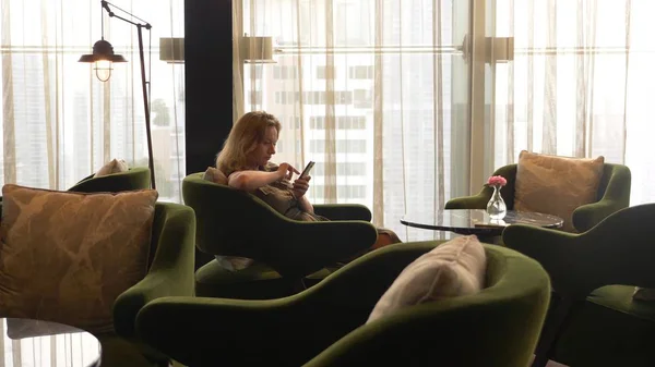 O blondă folosește telefonul într-o cafenea cu vedere de la fereastră la zgârie-nori . — Fotografie, imagine de stoc