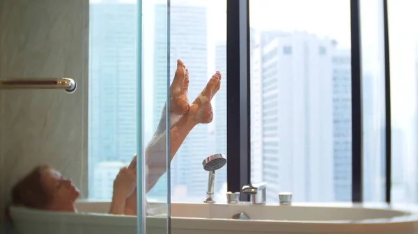 Schöne Frau genießt entspannendes Bad im luxuriösen Badezimmer mit Fenster. Lifestyle- und Beauty-Care-Konzept. Blick aus dem Fenster auf die Wolkenkratzer. — Stockfoto