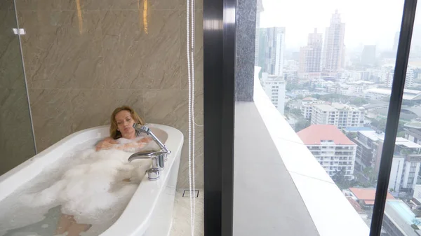 Mulher bonita desfrutando de banho relaxante em banheiro de luxo com uma janela. Estilo de vida e conceito de cuidados de beleza. vista da janela para os arranha-céus . — Fotografia de Stock