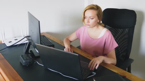 Hermosa mujer joven que trabaja en un ordenador portátil y un ordenador mientras está sentado en un escritorio . — Vídeos de Stock