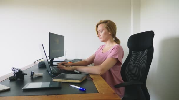 Hermosa mujer joven que trabaja en un ordenador portátil y un ordenador mientras está sentado en un escritorio . — Vídeo de stock