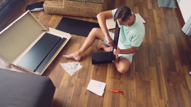 En ung man samlar självständigt möbler i vardagsrummet i sitt hus. En man samlar ett databord. — Stockvideo