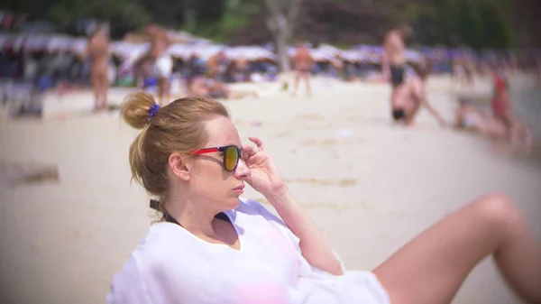 Una mujer pensativa y triste, se sienta sola en la arena en una playa llena de gente, borrosa de fondo — Foto de Stock