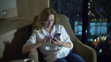 Genç, güzel sarışın kadın geceleri gökdelenler bakan panoramik pencereli bir odada bir sandalyeye bir smartphone kullanarak. arka plan bulanıklık.
