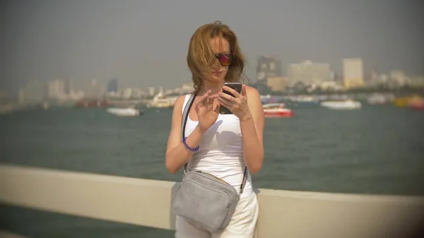 Vrouw in zonnebril, gebruikt haar smartphone terwijl staande op de pier. — Stockfoto