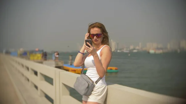 Женщина в солнечных очках, использует свой смартфон, стоя на пирсе . — стоковое фото