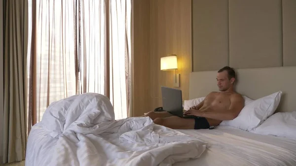 Молодой человек сидит в постели со своим ноутбуком. Он работает над этим. . — стоковое фото
