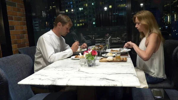 愉快的夫妇说话和吃饭在酒吧俯瞰摩天大楼, 在晚上, 模糊的背景 — 图库照片