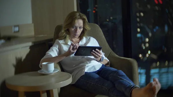 Joven, hermosa mujer rubia usando un teléfono inteligente, en una silla en una habitación con una ventana panorámica con vistas a los rascacielos por la noche. desdibujar el fondo . — Foto de Stock