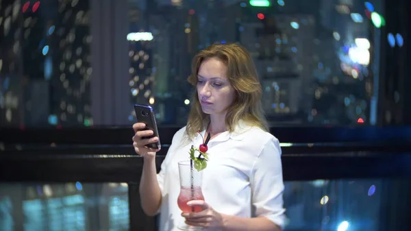 Молодая блондинка расслабляется и пьет коктейль в баре с видом на небоскребы ночью. Фоновое размытие — стоковое фото