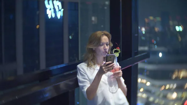 stock image Young woman blonde relaxing and drinking cocktail in bar with a view of the skyscrapers at night. background blur