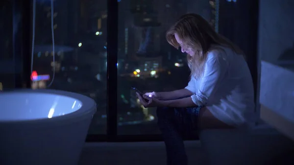 En kvinna som sitter på toaletten i en lyxig toalett med ett panoramafönster på natten med en telefon och arbetar. — Stockfoto