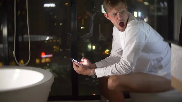 Un hombre sentado en el inodoro en un lujoso baño con una ventana panorámica por la noche con un teléfono y trabajando . — Foto de Stock