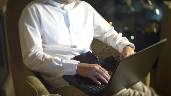 Un jeune et beau homme utilisant un ordinateur portable sur une chaise dans une chambre avec une fenêtre panoramique donnant sur les gratte-ciel la nuit. brouiller le fond . — Photo