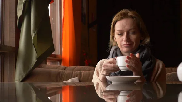 Mujer joven bebiendo té en la cafetería cerca de la ventana con vista al mar. una mujer en una bufanda y suéter está bebiendo té y tomando el sol en un café . — Foto de Stock