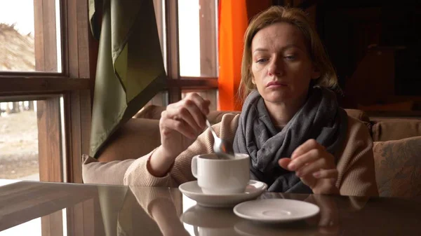 Jovem mulher bebendo chá no café perto da janela com vista para o mar. uma mulher em um cachecol e suéter está bebendo chá e se saciando em um café . — Fotografia de Stock