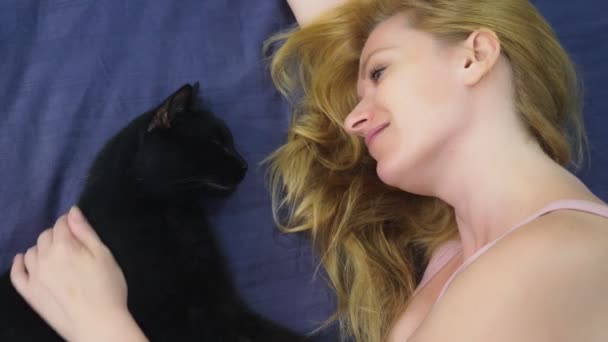 고양이와 소녀입니다. 아름 다운 금발 소녀 침대에 누워 하 고 그녀의 검은 고양이 애 무. 동물에 대 한 배려. 애완 동물에 대 한 사랑 — 비디오