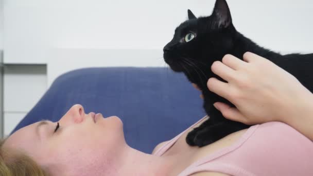 Une fille avec un chat. Belle fille blonde allongée au lit et caressant son chat noir. Prendre soin des animaux. amour pour les animaux — Video