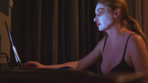 Νεαρή γυναίκα με καλλυντικά μάσκα στο πρόσωπο που εργάζεται με φορητό υπολογιστή στο σπίτι το βράδυ. φυσικότητα — Αρχείο Βίντεο