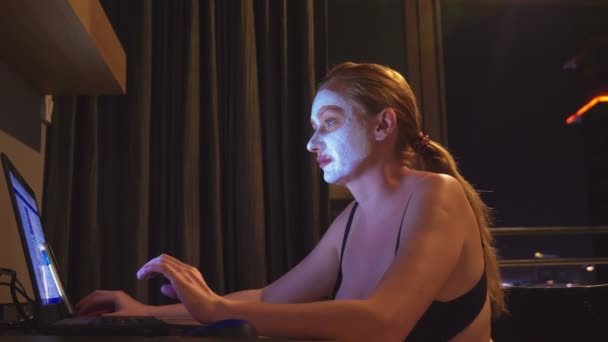 Ung kvinna med kosmetiska mask på ansikte arbeta med laptop hemma på kvällen. naturlighet — Stockvideo