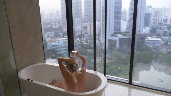 窓のある豪華なバスルームで髪を洗う美しい女。生命および美の方法の概念。高層ビルの窓からの眺め — ストック写真
