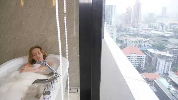 Hermosa mujer disfrutando de un baño relajante en un baño de lujo con ventana. Concepto de estilo de vida y cuidado de belleza. vista desde la ventana a los rascacielos . — Foto de Stock