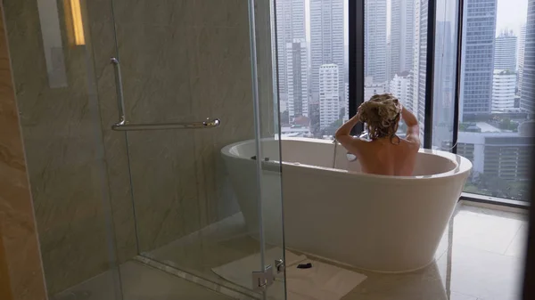 Schöne Frau wäscht ihre Haare in einem luxuriösen Badezimmer mit Fenster. das Konzept einer Lebensweise und Schönheit. Blick aus dem Fenster auf die Wolkenkratzer — Stockfoto