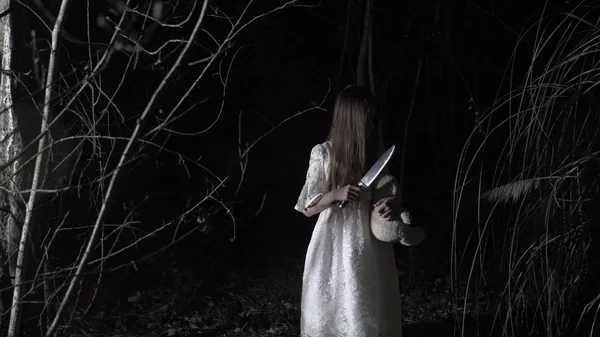 Una niña fantasma con el pelo largo y negro, en blanco, vagando por el bosque con un cuchillo y un juguete suave . — Foto de Stock