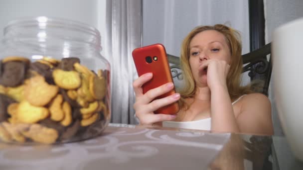Голодна сумна жінка сидить у вітальні за столом вночі, вона їсть печінку і використовує свій смартфон — стокове відео