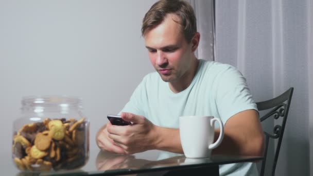 Hombre triste hambriento se sienta en una sala de estar en una mesa por la noche, se come un hígado y utiliza su teléfono inteligente — Vídeos de Stock
