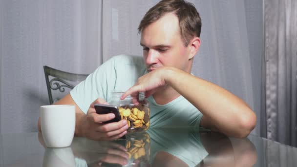 Hombre triste hambriento se sienta en una sala de estar en una mesa por la noche, se come un hígado y utiliza su teléfono inteligente — Vídeos de Stock