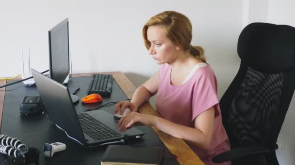 Gyönyörű fiatal nő dolgozik a laptop és a számítógép egy íróasztalnál ülve. — Stock videók