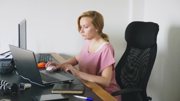 Bela jovem mulher trabalhando em um laptop e computador enquanto sentado em uma mesa . — Vídeo de Stock