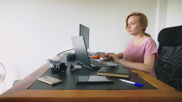 Hermosa mujer joven que trabaja en un ordenador portátil y un ordenador mientras está sentado en un escritorio . — Vídeos de Stock